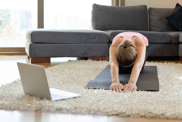 Online άθληση προπόνηση γιόγκα. νεαρή γυναίκα και κάνει ασκήσεις στο στρώμα γιόγκα απέναντι από το laptop με online master class. εκπαίδευση στο σπίτι. - Φωτογραφία, εικόνα