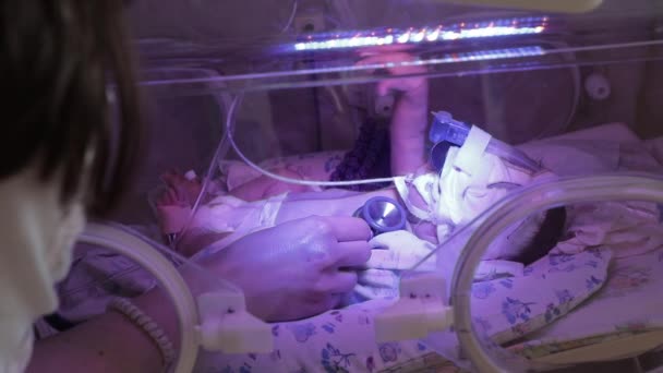 Ženský doktor protlačí její ruku dírou v inkubátoru a poslouchá tlukot srdce dítěte stetoskopem. Dítě se pohne, doktor opraví trubičku připojenou k ruce novorozence. Zavřít - Záběry, video