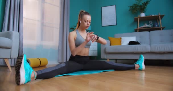 Young fit woman warm up stratching on the floor using app on smart watch to check heartbeat before sport workout for healthy lifestyle at home. Sport and fitness. Training and wellness concept. - Video
