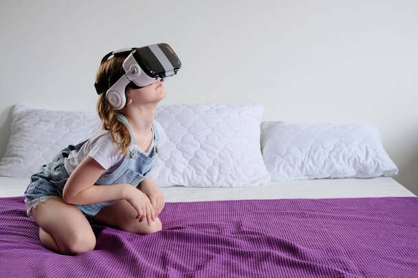 Games of a little girl in glasses of venrtualnoy reality. Sitting at home on the couch, the girl got into another world through virtual glasses - Foto, imagen