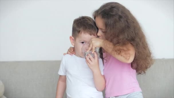 Pretty sister, a nanny with a younger brother are sitting on the couch, a loving happy sister takes care of her little brother, gives a mug of water, entertains the child. - Záběry, video