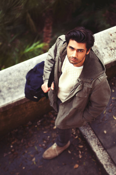 Handsome young italian man walking in the street. Seen from above. Stylish hair and coat outdoors wearing winter jackets, shoulder bag and autumn clothes. Outdoors in a natural park in Rome, Italy. - Photo, image