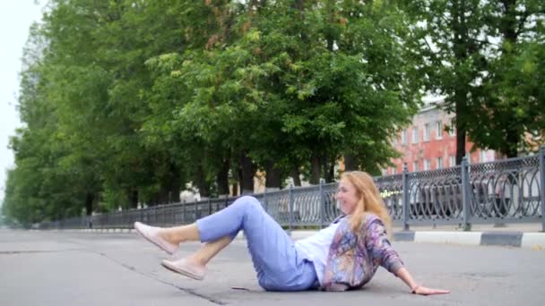 Drunk young girl with long blond red hair in blue trousers, sitting on her ass on the roadway in the city early in the morning, swinging in different directions, raising her legs, smiling. Crazygirl. - Video