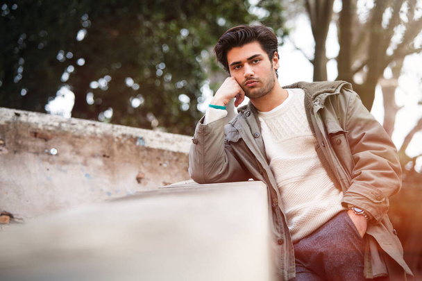Модный молодой человек позирует на улице, опираясь на стену с покоящимся локтем. Зимняя одежда с пальто и белым свитером. Зеленые деревья позади
. - Фото, изображение