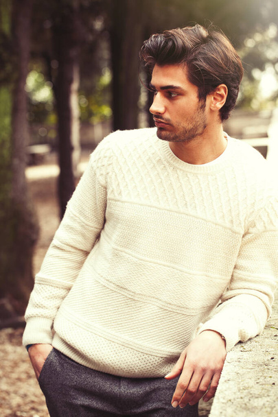 Handsome serene and pensive young man model in a trees park. Charming young and handsome man with stylish hair and white sweater. Outdoors in a park with a series of trees. Peaceful and calm attitude. - Foto, Bild