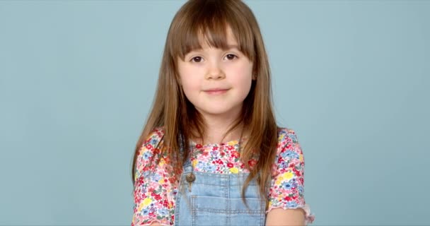 Süßes kleines Mädchen 6-7 Jahre alt posiert in Latzhose und Bluse mit Blumenmuster auf blau - Filmmaterial, Video