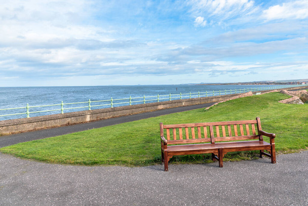 Tyhjä puinen penkki rauhallisessa merenrantapuistossa osittain pilvisenä syyspäivänä. Musselburgh, Skotlanti, Yhdistynyt kuningaskunta
. - Valokuva, kuva