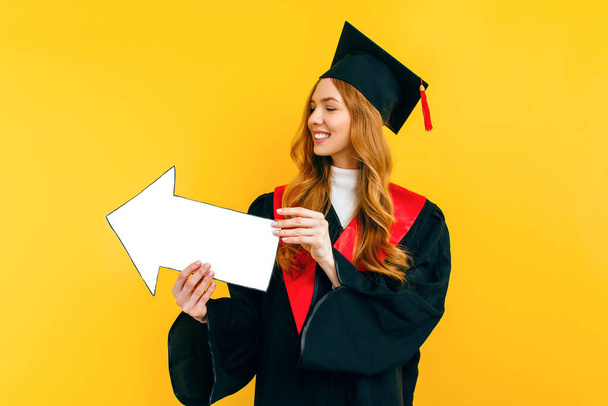 A happy, beautiful graduate holds a clean paper layout, pointing to a copy of the space on a yellow background - Fotografie, Obrázek