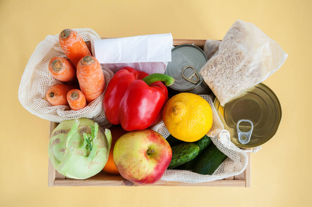 Donation box with various food. Canned food, cereals, fruits, vegetables. Yellow background, flat lay - Фото, зображення