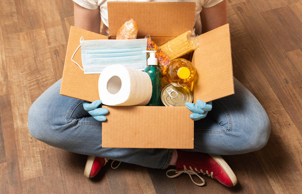 Quarantine food delivery service for those who need. A person sitting on a floor and holding donation box with food, toilet paper and sanitizer. Stay home during a COVID-19. High quality photo - Photo, Image