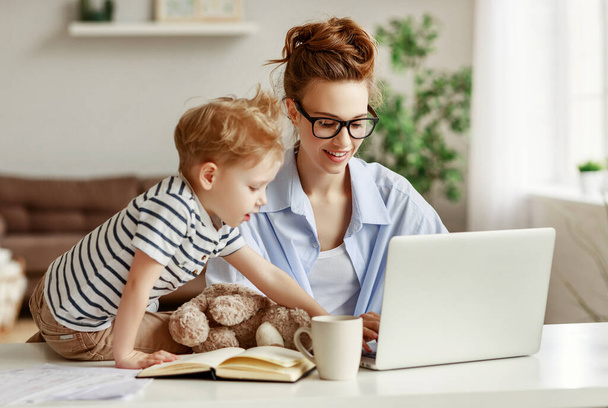 Cheerful female entrepreneur and little boy smiling and typing on laptop keyboard while sitting at table and working on freelance project at home togethe - Photo, Image