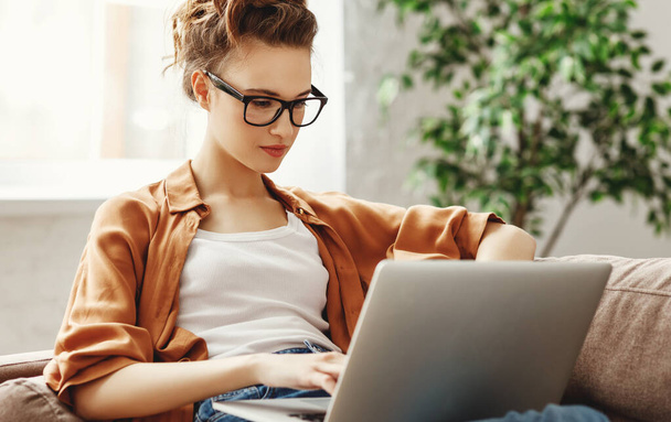 Slimme jonge vrouw met bril zittend op de bank en freelance project op laptop in gezellige woonkamer thuis - Foto, afbeelding