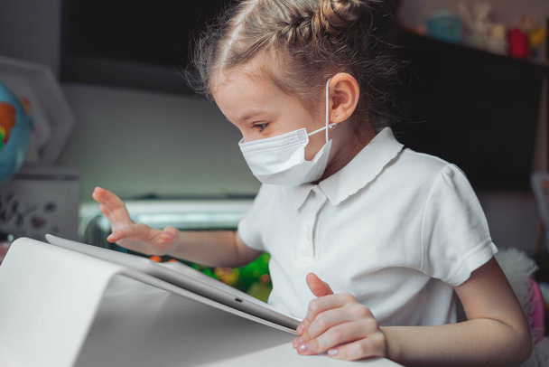 Caucasian preteen girl with medical mask on her face concentrated on task with tablet. Concept of distance learning in isolation while coronavirus - Zdjęcie, obraz