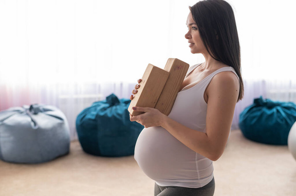 Caucasian pregnant woman with equipment holds pilates equipment in the hall with chair bags. Expectant mother with blocks for yoga. - Foto, imagen
