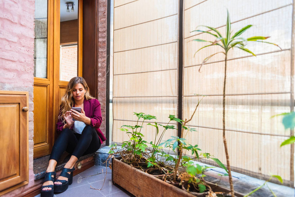 élégante jeune femme appel vidéo avec téléphone mobile, assis sur le sol avec des plantes dans sa maison
. - Photo, image