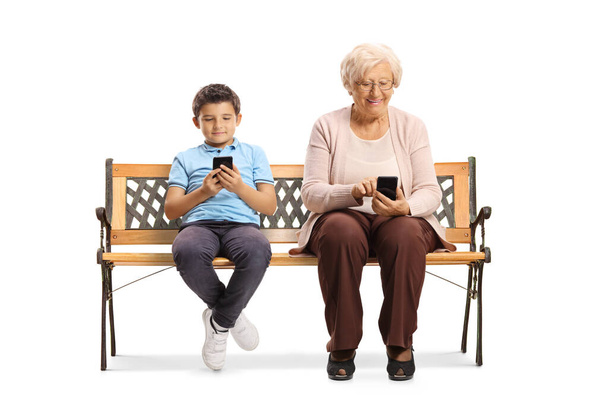 Yaşlı bir kadın ve bir çocuk bankta oturuyor ve beyaz arka planda izole edilmiş cep telefonları kullanıyorlar.  - Fotoğraf, Görsel