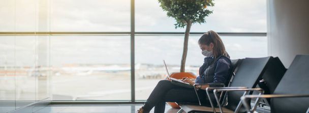 Egy nő ül a reptéren egy laptoppal orvosi maszkban. Egy fiatal lány várja az indulást a kapunál, online dolgozik. Társadalmi felelősségvállalás, vírusvédelem. - Fotó, kép