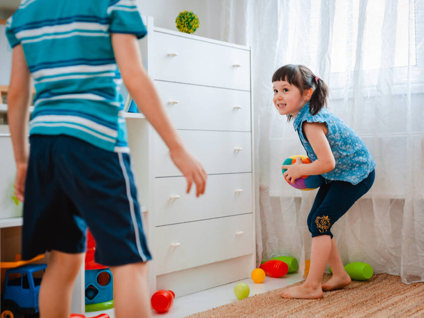 ντόπια παιδιά αγόρι και ένα κορίτσι παίζουν σε ένα παιδικό δωμάτιο παιχνιδιού, ρίχνοντας μπάλα. έννοια της αλληλεπίδρασης αδέλφια, επικοινωνία, αμοιβαίο παιχνίδι, καραντίνα, αυτο-απομόνωση σπίτι, αδελφός αδελφή. - Φωτογραφία, εικόνα