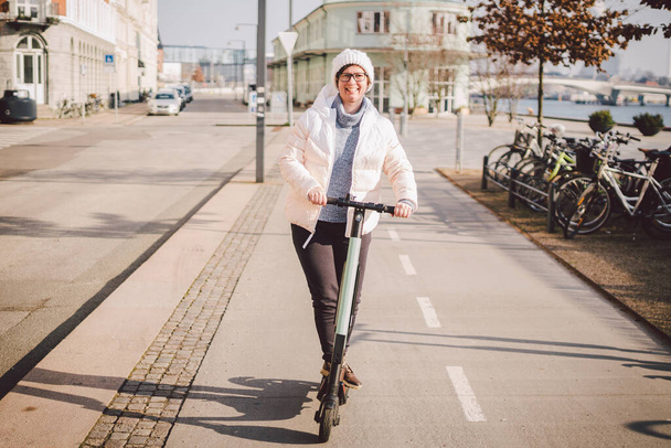 女性は電動スクーターを借りており、コペンハーゲンのデンマークで晴れた冬の日に乗る予定です。生態学的にきれいな輸送のテーマ。都市交通電動スケートボードの新しいクイックビュー. - 写真・画像