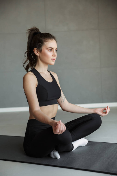 Photo of focused young woman in sportswear meditating while sitting with legs crossed on mat - Zdjęcie, obraz
