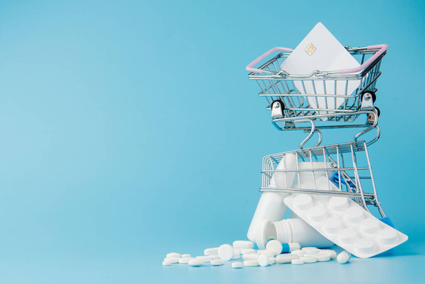 scattered variety pills, drugs, spay, bottles, thermometer, syringe and empty shopping trolley cart on blue background. pharmacy shopping concept. - Photo, Image