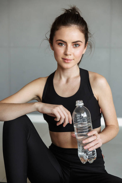 Photo of pleased brunette woman in sportswear drinking water while working out at gym - Photo, Image