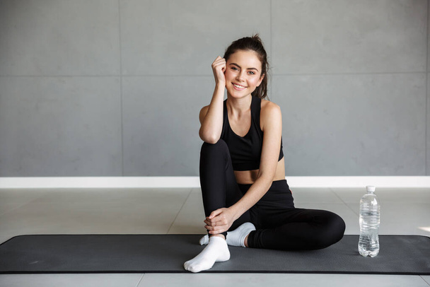Photo of pleased brunette woman in sportswear smiling while sitting with water bottle on mat - Fotó, kép