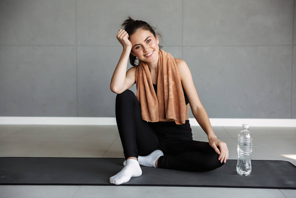 Photo of joyful brunette woman in sportswear smiling while sitting with water bottle and towel on mat - Fotografie, Obrázek