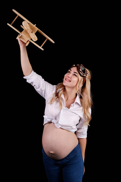Νεαρή έγκυος όμορφη κοπέλα με γυαλιά πιλότου και με ένα αεροπλάνο στο χέρι. απομονωμένο σε μαύρο φόντο - Φωτογραφία, εικόνα