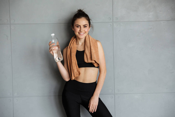 Photo of smiling nice woman in sportswear posing with towel over her neck and water bottle near wall - Fotoğraf, Görsel