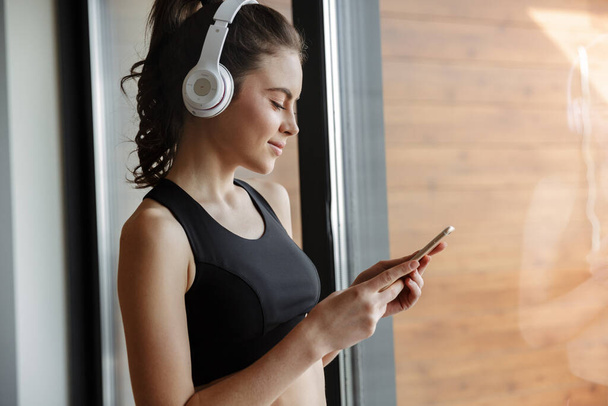Photo of pleased seductive woman in sportswear using headphones and smartphone while standing near window - Фото, изображение