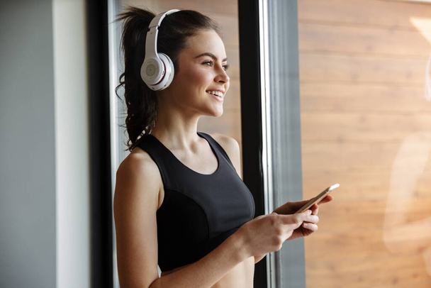 Photo of smiling seductive woman in sportswear using headphones and smartphone while standing near window - 写真・画像