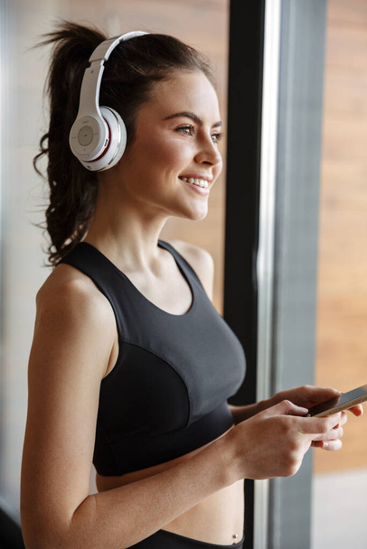 Photo of smiling seductive woman in sportswear using headphones and smartphone while standing near window - Photo, image