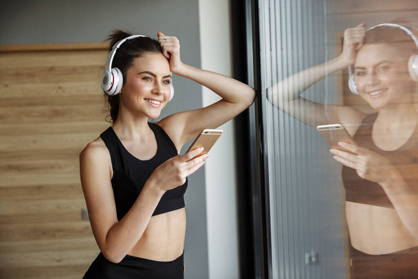 Photo of smiling seductive woman in sportswear using headphones and smartphone while leaning on window - Фото, изображение