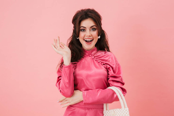 Portrait of a beautiful cheerful young brunette woman with bright makeup wearing fashionable clothes standing isolated over pink background, holding a purse, posing - Photo, Image