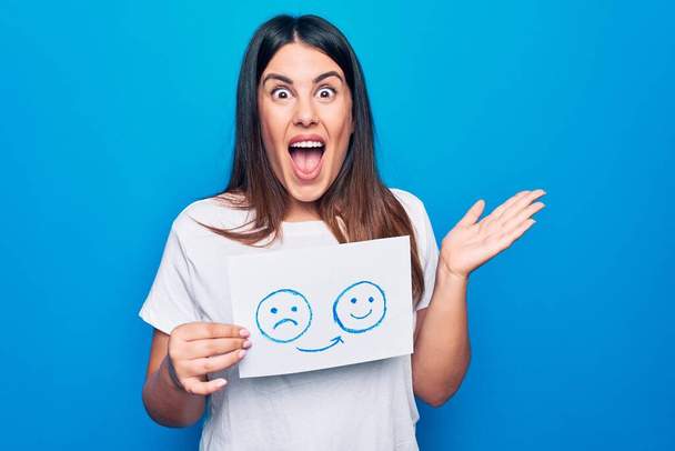 Woman asking for psychology change holding paper with unhappy to happy emotion message celebrating achievement with happy smile and winner expression with raised hand - Photo, Image