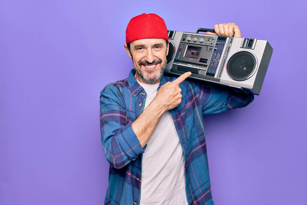Hombre de mediana edad con gorra escuchando boombox vintage sobre fondo púrpura aislado sonriendo feliz señalando con la mano y el dedo
 - Foto, imagen