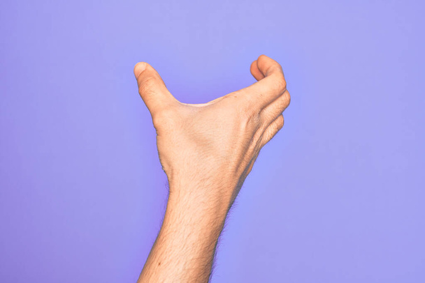 Hand eines kaukasischen jungen Mannes mit Fingern über isoliertem lila Hintergrund, der unsichtbare Gegenstände hält, leere Hand beim Ausschneiden und Greifen - Foto, Bild