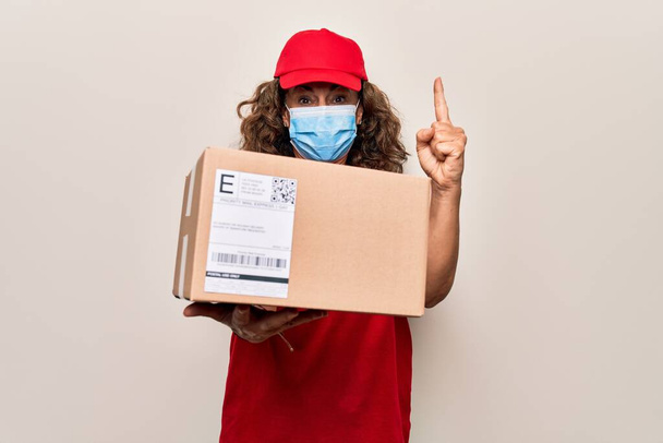 Orta yaşlı teslimatçı kadın Coronavirus koruma maskesi takıyor ve elinde bir fikir ya da soruyla gülümseyen karton bir paket tutuyor. - Fotoğraf, Görsel