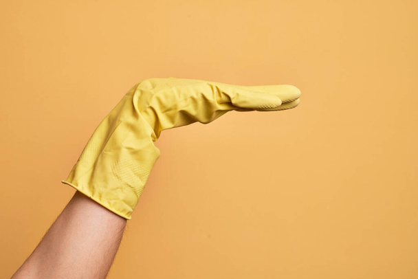 Hand of caucasian young man with cleaning glove over isolated yellow background with flat palm presenting product, offer and giving gesture, blank copy space - Фото, изображение