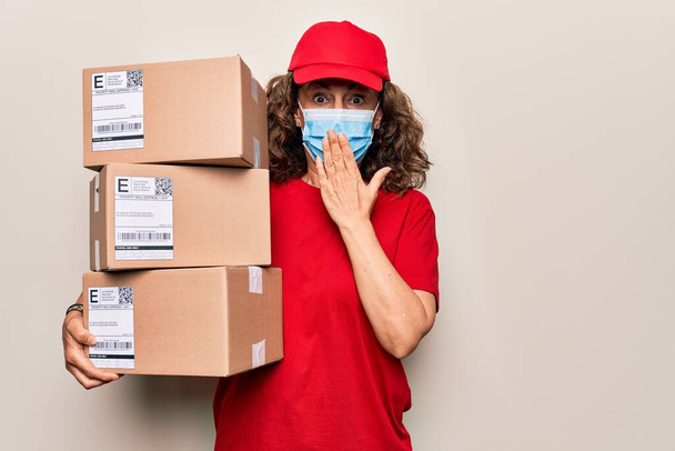 Coronavirus koruma maskesi takan orta yaşlı bir teslimatçı kadın elinde karton kutular, ağzı elle kapatıyor, şok geçiriyor ve hata yapmaktan korkuyor. Şaşırtıcı ifade. - Fotoğraf, Görsel