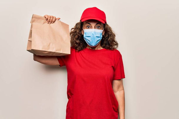 Középkorú futár nő visel koronavírus védelmi maszk és a gazdaság papírzacskó rémült és csodálkozott nyitott szájjal meglepetés, hitetlenség arc - Fotó, kép