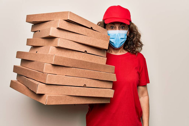 Žena ve středním věku nosí koronavirovou ochrannou masku a drží kartónové krabice od pizzy vyděšené a ohromený s otevřenými ústy pro překvapení, nevěřící tvář - Fotografie, Obrázek