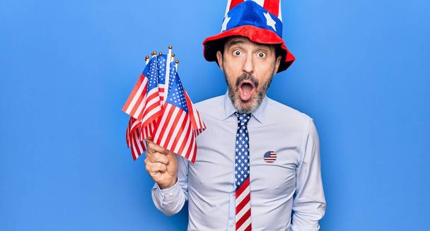 Middle age handsome patriotic man wearing united states hat and tie holding american flags scared and amazed with open mouth for surprise, disbelief face - Φωτογραφία, εικόνα