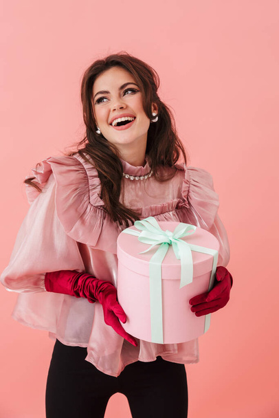 Portrait of a beautiful cheerful young brunette woman with bright makeup wearing fashionable clothes standing isolated over pink background, holding present box - Photo, Image