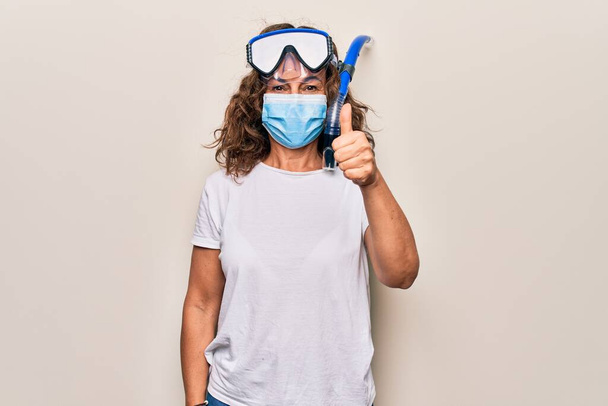 Middle age woman wearing coronavirus protection mask and diving gogles glasses as canceled vacation smiling happy and positive, thumb up doing excellent and approval sign - Foto, Imagem