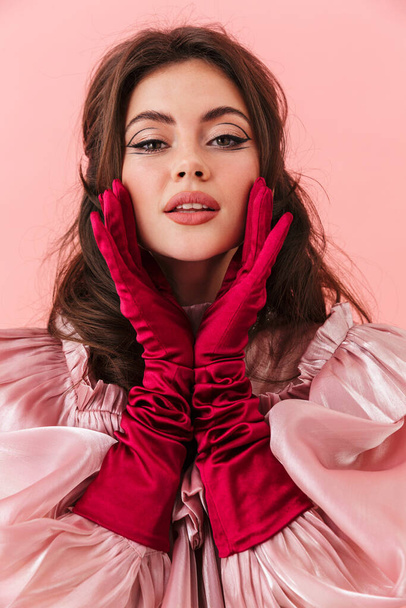 Portrait of a beautiful young brunette woman with bright makeup wearing fashionable clothes standing isolated over pink background, posing - Фото, зображення