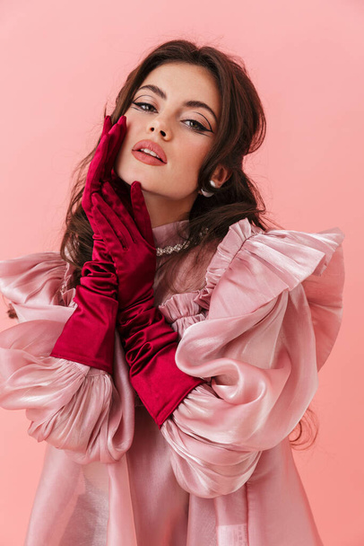 Portrait of a beautiful young brunette woman with bright makeup wearing fashionable clothes standing isolated over pink background, posing - Foto, afbeelding