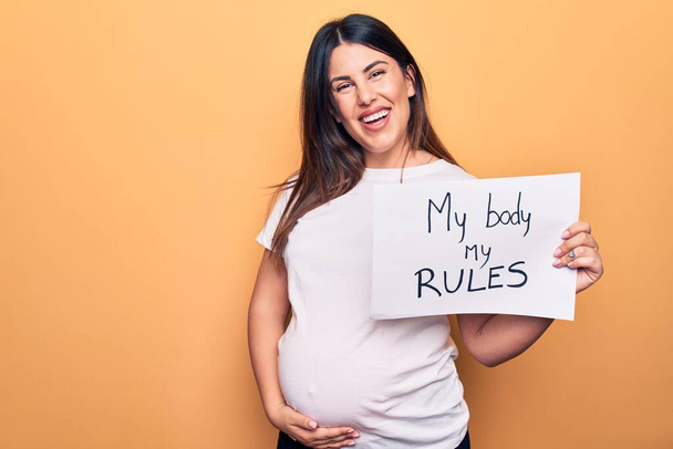 Молода красива брюнетка вагітна жінка тримає прапор з моїм тілом мої правила повідомлення виглядає позитивним і щасливим стоячи і посміхаючись з впевненою посмішкою показуючи зуби
 - Фото, зображення