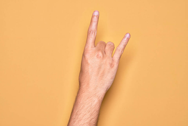 ręka kaukaski młody człowiek pokazując palce na odizolowanym żółtym tle gestykulując rock and roll symbol, pokazując obsceniczne rogi gest - Zdjęcie, obraz